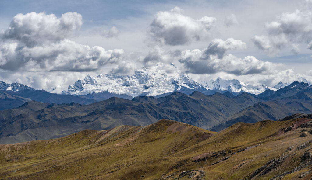 Snow-capped mountain peaks with rugged terrain and rolling green hills under cloudy sky.